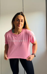 A girl wearing a cropped t-shirt .  The shirt is a female git and has a round neck and standard sleeve length.  The shirt has The Defiant Co logo across the centre of the chest.  The shirt is pink in colour.