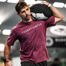 Load image into Gallery viewer, A photo showing a guy wearing a slightly oversized Acid Wash unisex T-Shirt while carrying sandbag during a workout.  The shirt has the famous ‘The Defiant Co’ logo across the front of the chest.  The acid Wash effect of the shirt means no two are alike and provides the perfect blend of fashion and defiance. The shirt is burgundy with a slightly black wash.