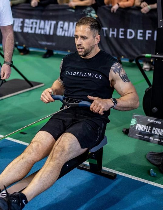 The photo shows a guy wearing The Defiant Co sleeveless t-shirt whilst rowing during a CrossFit workout.  The sleeveless is black and has the famous The Defiant Co logo across the front of the chest in white. The guy is an athlete competing at The Defiant Games.