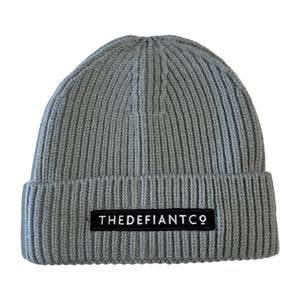 A photo showing a beanie hat. The hat has a ribbed finish with a roll cuff.  The cuff has The Defiant Co logo embroidered on the centre of the front in white on a black background. The beanie hat is light grey.