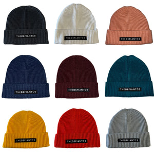 A collage showing a selection of beanie hats in a variety of colours. The hats have a ribbed finish with a roll cuff.  The cuff has The Defiant Co logo embroidered on the centre of the front in white on a black background. 