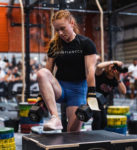 A girl performing dummbbell box step overs during a crossfit competition. She is wearing a cropped t-shirt from The Defiant Co displaying the company logo across the centre of the chest.