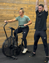 Load image into Gallery viewer, A girl and a guy in the gym wearing the amazing DFNT. Performance Joggers.  The girl sits on and Assault Bike while the guy performs a kettle bell swing.  The joggers have a DFNT. logo min raised rubber that sits on the left quad and The Defiant Co screen printed in a subtle grey tracking down the left lower shin.  The joggers have two zip pockets on either leg and have an elasticated and draw string waistband.
