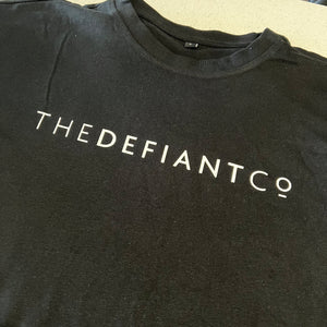 A photo showing an oversized The Defiant Co T-Shirt.  The shirt has the famous ‘The Defiant Co’ logo across the front of the chest.  The shirt has a round neck and is oversized.  This particular version is black.