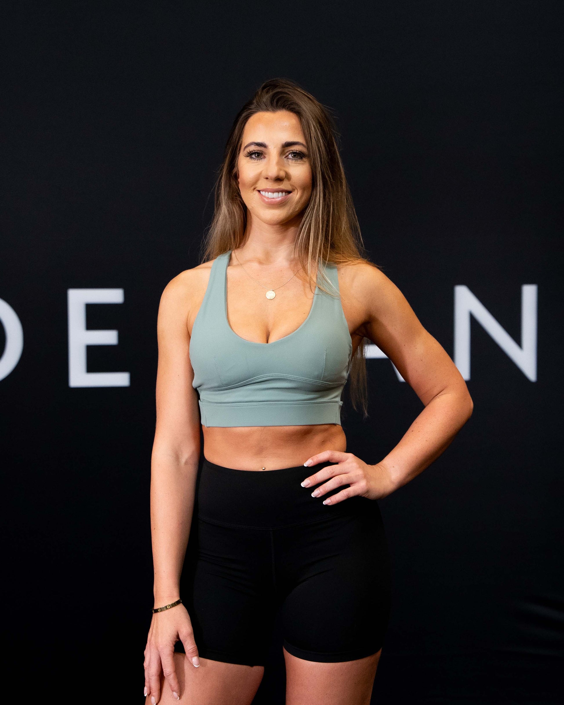 A girl against a black The Defiant Co backdrop wearing the Infinity Sports bra with it's crossed back and supportive front.  The Defiant Co is criss-crossed over both straps across the back. The colour is light olive.