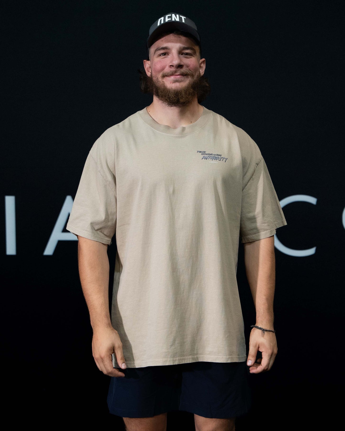 A guy against a black The Defiant Co backdrop wearing the faded bone proudly refusing to obey authority oversized tee and black performance shorts. 