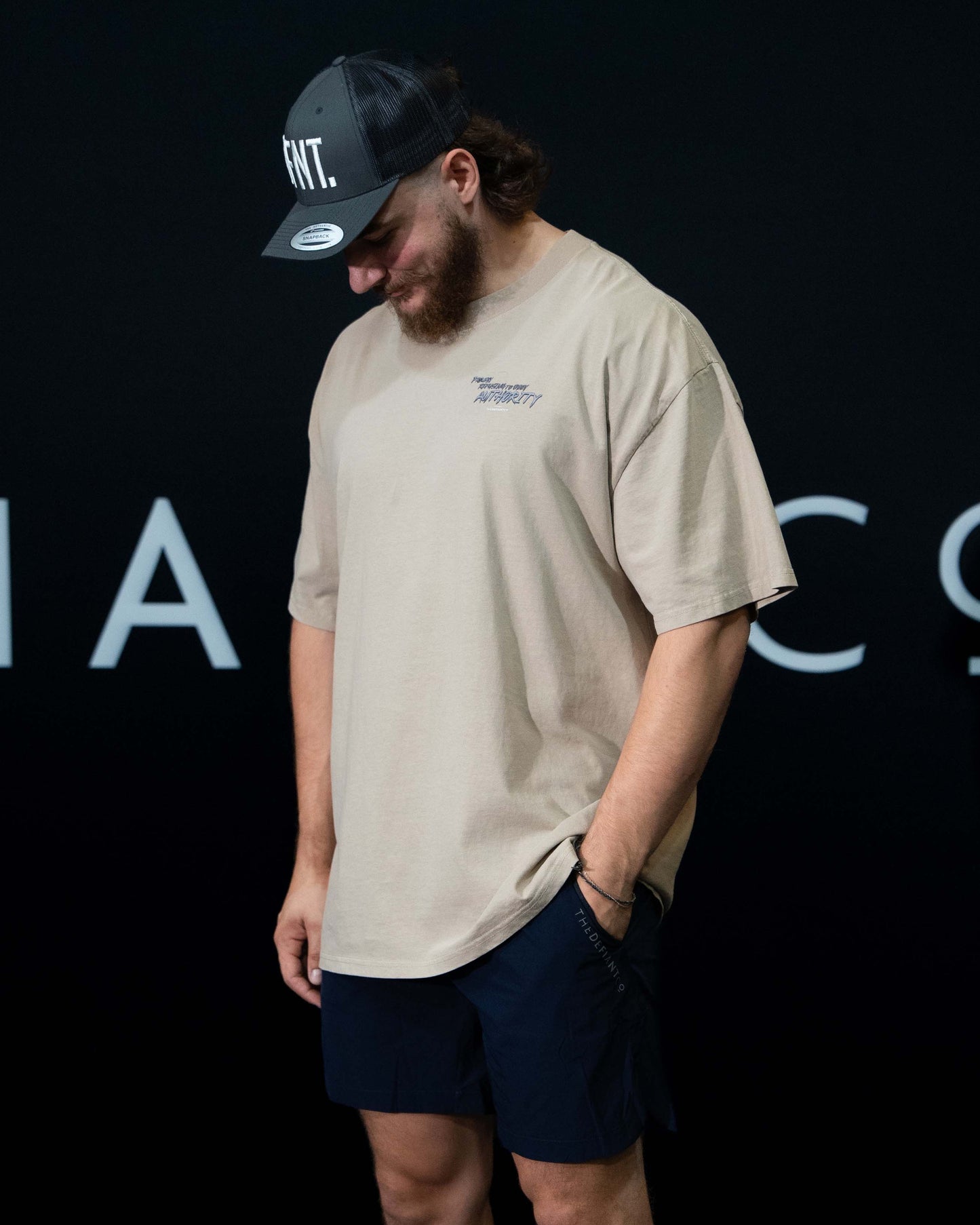 A guy against a black The Defiant Co backdrop wearing the faded bone proudly refusing to obey authority oversized tee and black performance shorts. 