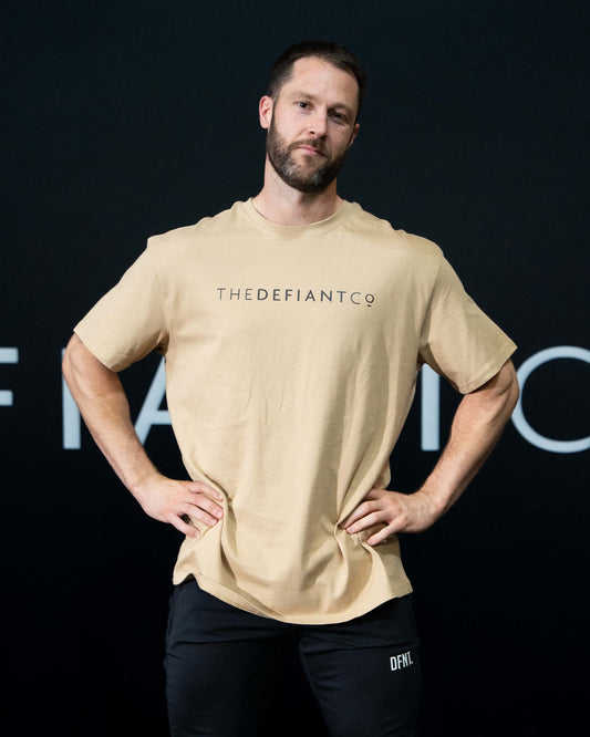 A guy against a black The Defiant Co backdrop wearing the oversized t-shirt.  The tee has the famous 'The Defiant Co' logo boldly displayed across the front. The colour is beige.