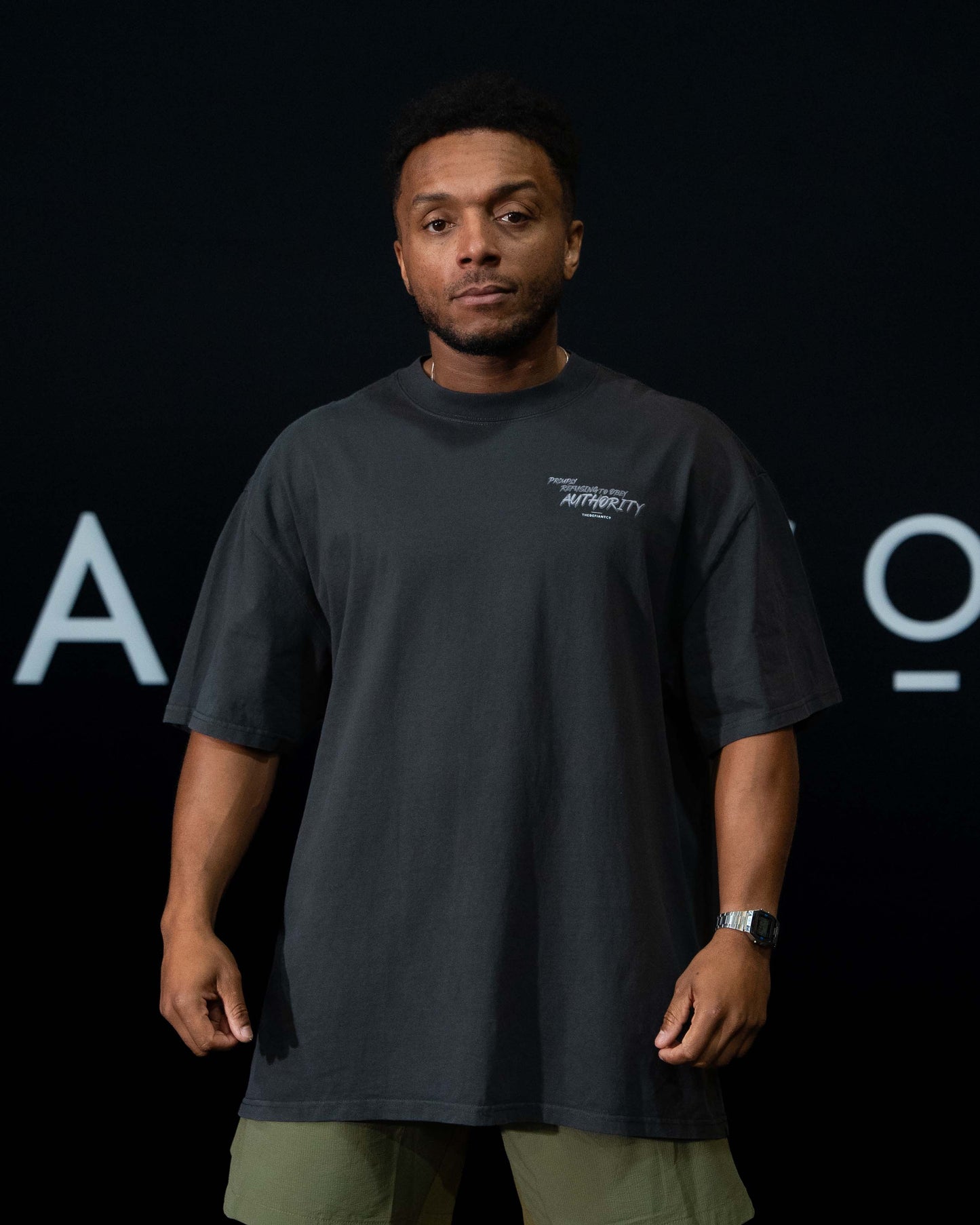 A guy against a black The Defiant Co backdrop wearing the faded black proudly refusing to obey authority oversized tee and olive green performance shorts. 