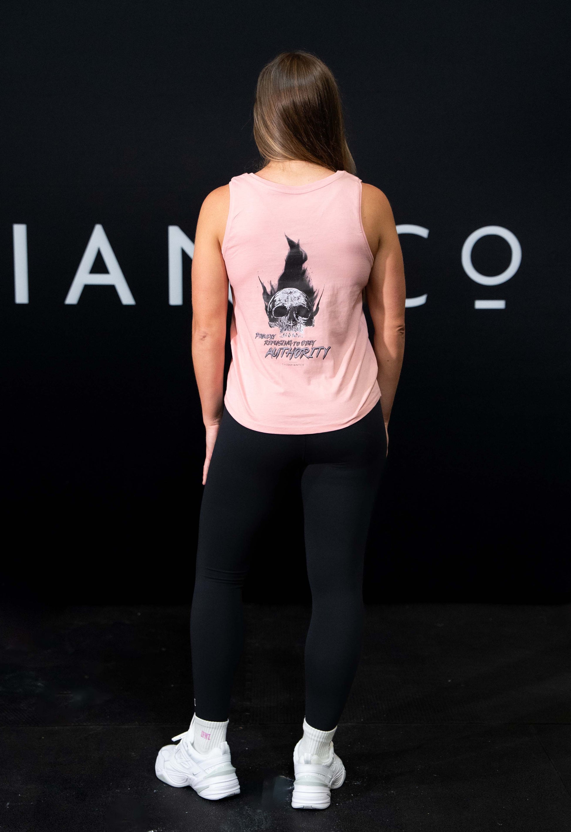 A girl against a black The Defiant Co backdrop wearing the Proudly Refusing to Obey Authority Vest Top.  The vest is slightly cropped with a high neckline.  It has a subtle left breast print and large skull and flames graphic on the back.  The colour is pink.