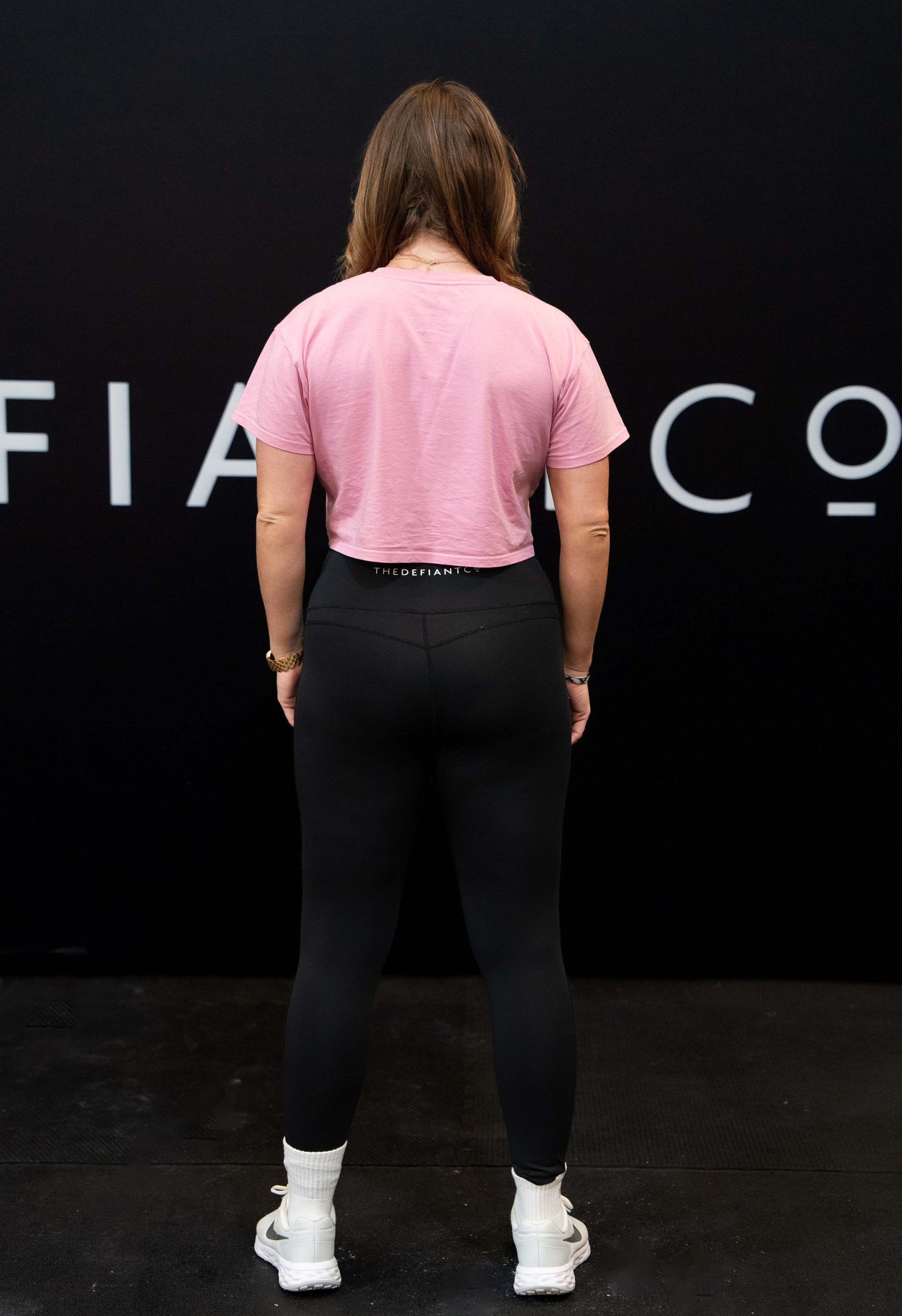 A girl against a black The Defiant Co backdrop wearing the TDCo cropped t-shirt in baby pink with the black performance leggings. 