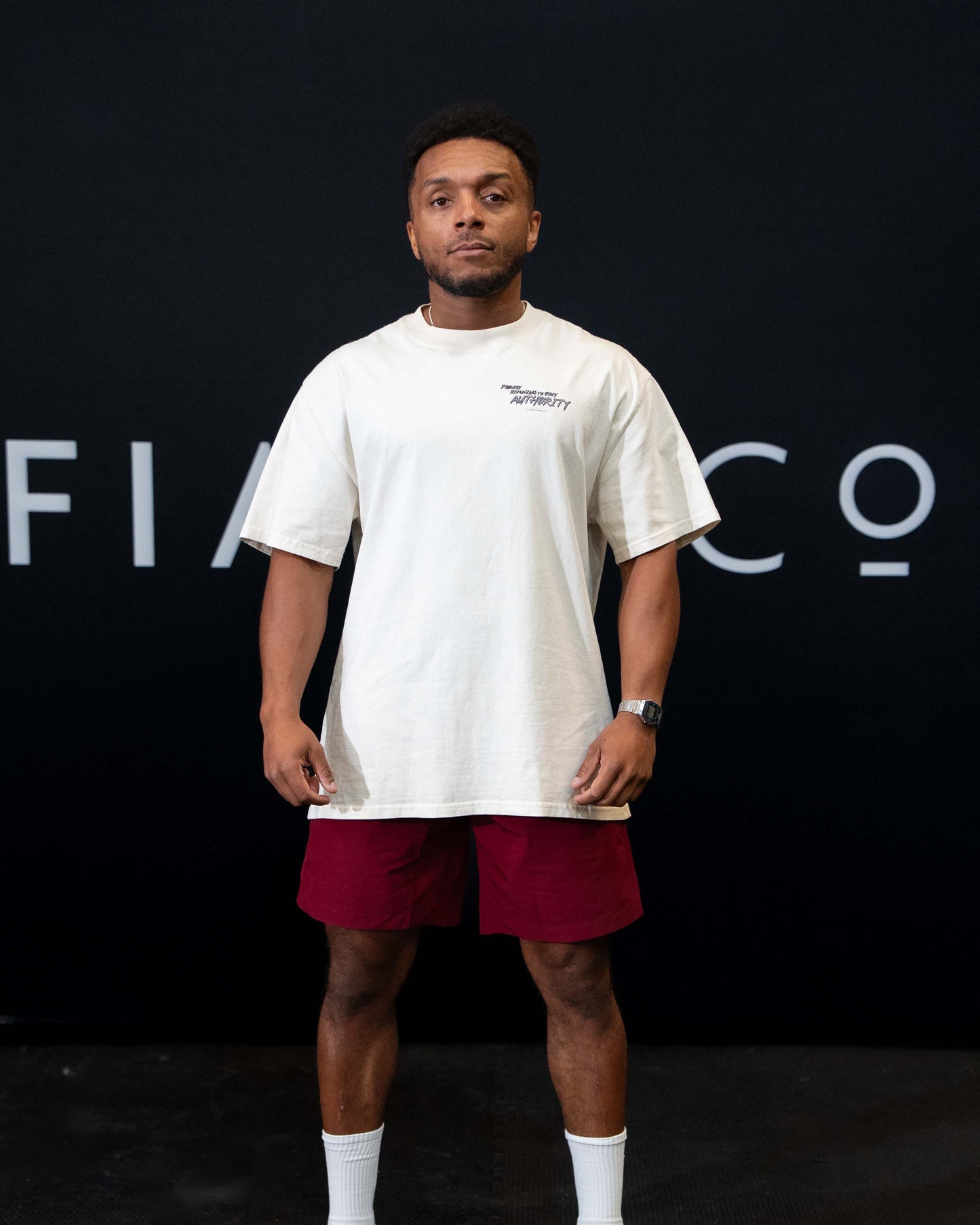 A guy against a black The Defiant Co backdrop wearing the faded ecru proudly refusing to obey authority oversized tee and burgundy performance shorts. 