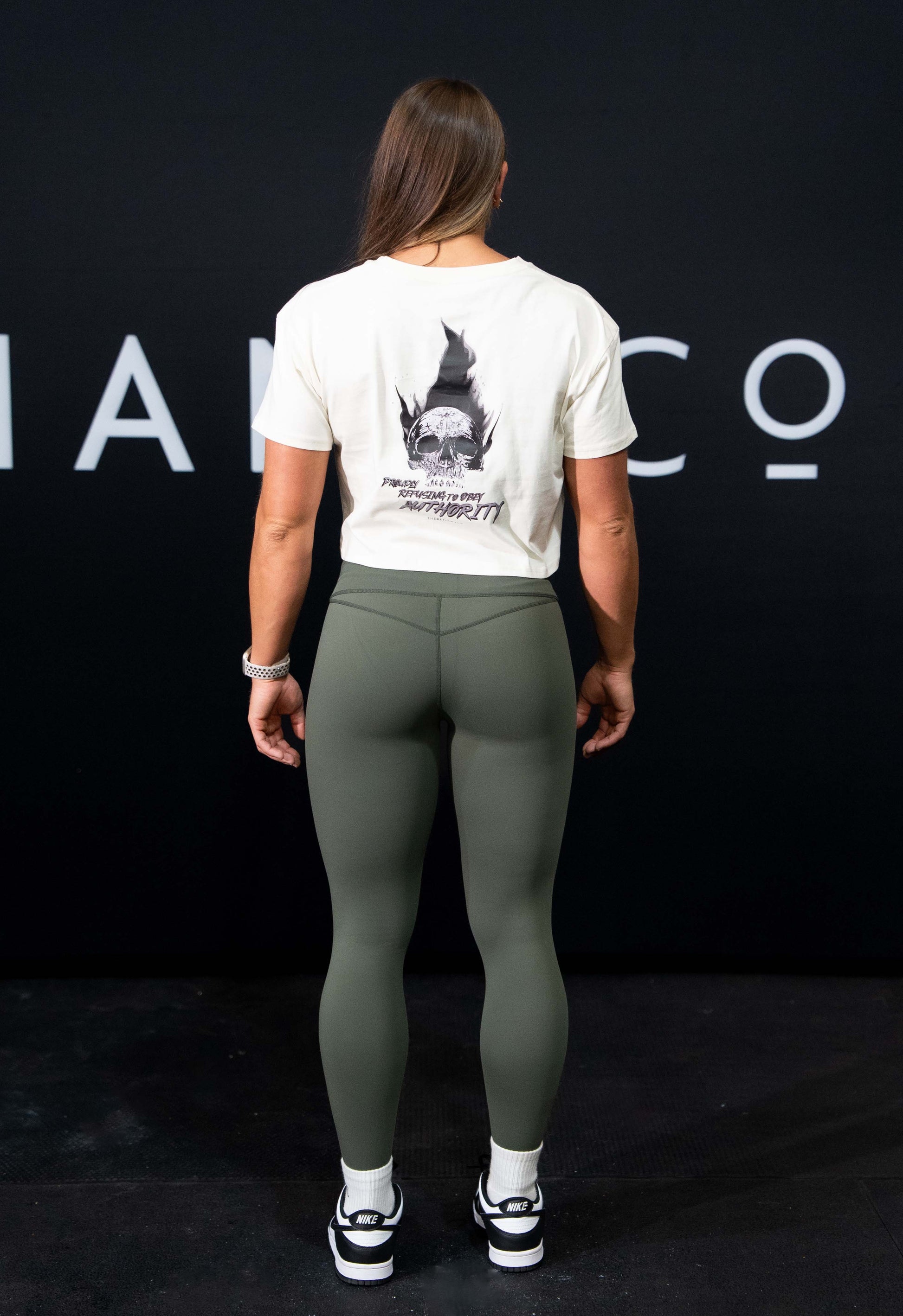 A girl against a black The Defiant Co backdrop wearing the proudly refusing to obey authority cropped t-shirt in ecru and the olive green TDCo performance leggings with white activewear socks and trainers. 