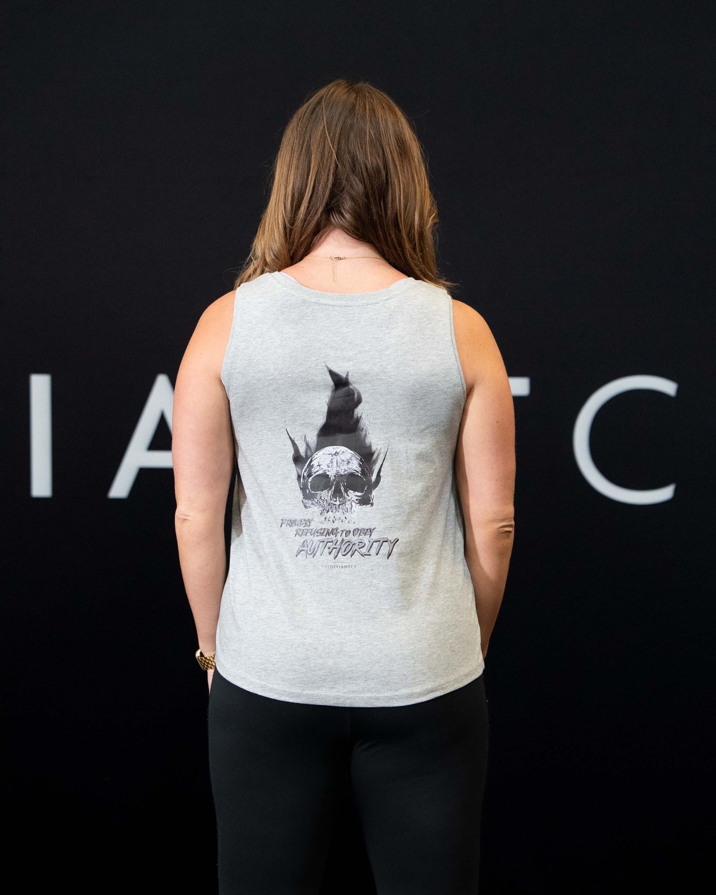 A girl against a black The Defiant Co backdrop wearing the Proudly Refusing to Obey Authority Vest Top.  The vest is slightly cropped with a high neckline.  It has a subtle left breast print and large skull and flames graphic on the back.  The colour is heather grey.