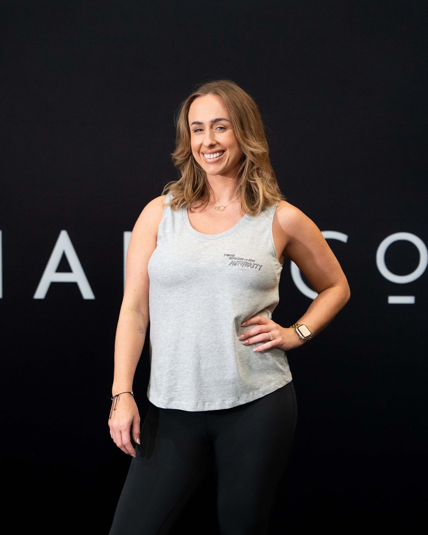 A girl against a black The Defiant Co backdrop wearing the Proudly Refusing to Obey Authority Vest Top.  The vest is slightly cropped with a high neckline.  It has a subtle left breast print and large skull and flames graphic on the back.  The colour is heather grey.