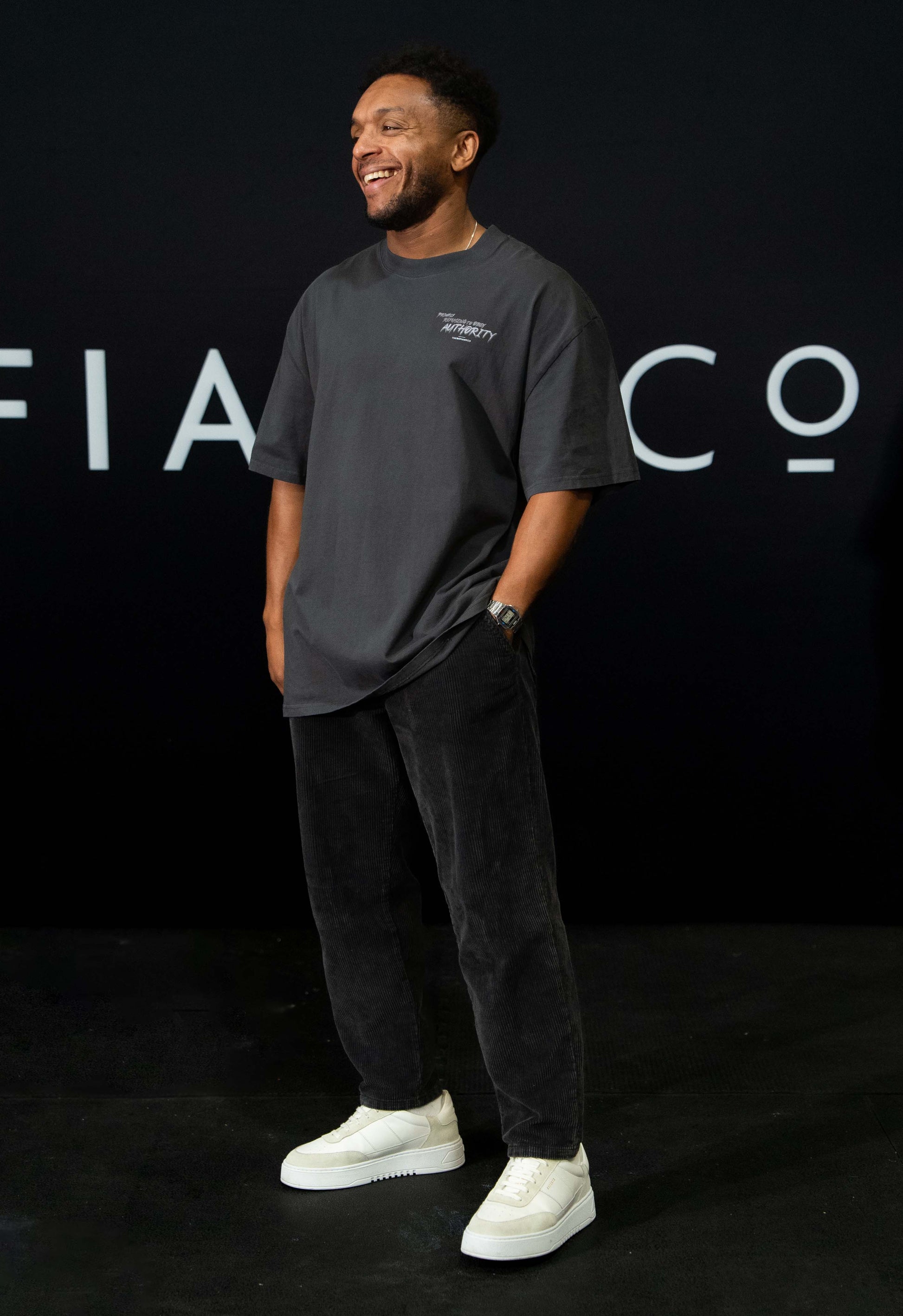 A guy against a black The Defiant Co backdrop wearing the faded black proudly refusing to obey authority oversized tee and black corduroy trousers. 