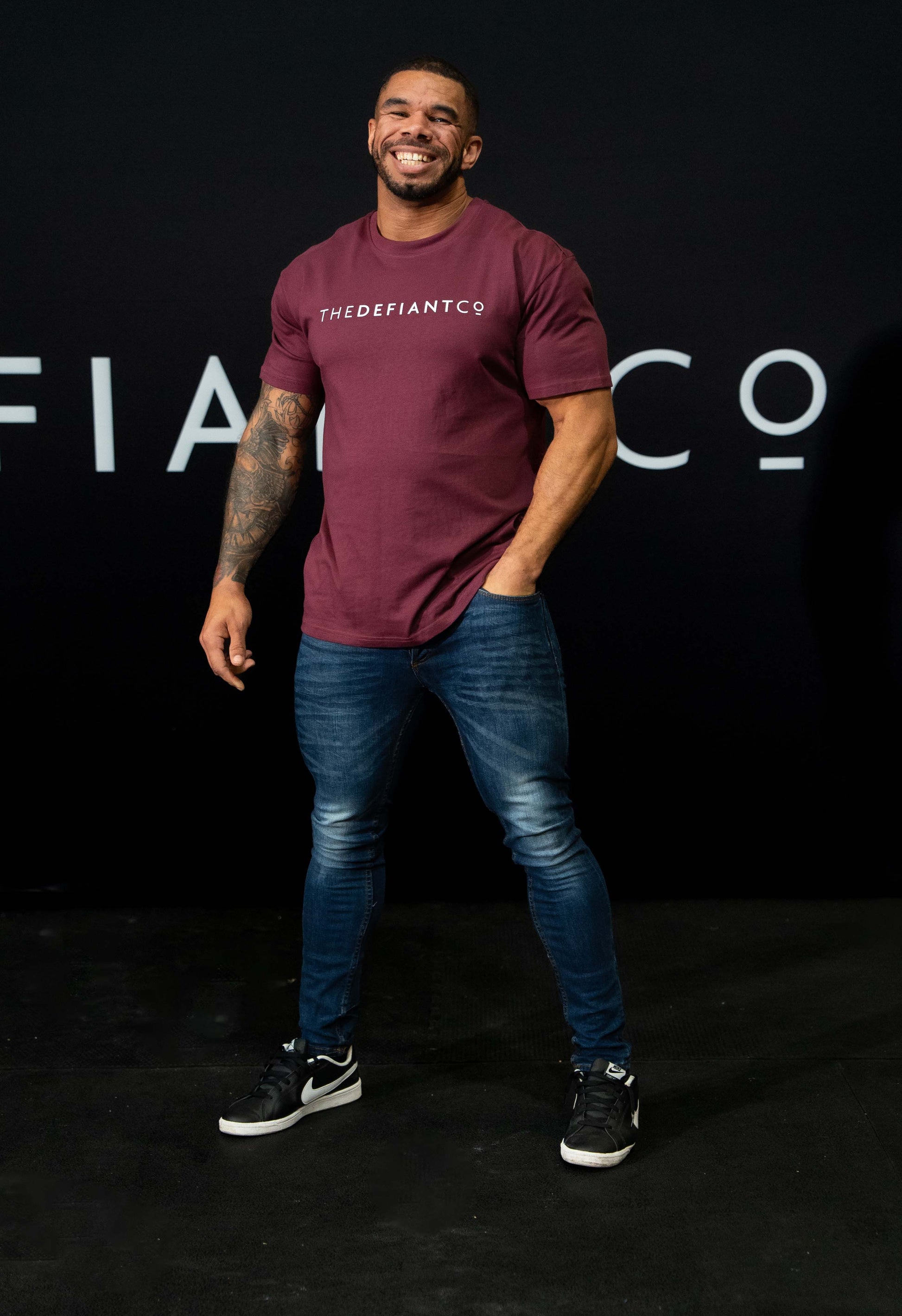 A guy against a black The Defiant Co backdrop wearing the oversized t-shirt.  The tee has the famous 'The Defiant Co' logo boldly displayed across the front. The colour is burgundy.