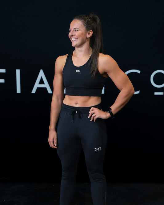 A girl against a black The Defiant Co backdrop wearing the DFNT. Black Jogging Bottoms and black sports bra.   It has a subtle DFNT. logo under the pocket. It has a drawstring waist and the pockets have zips. 