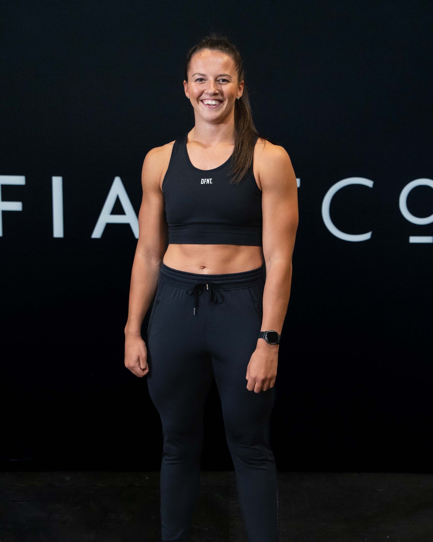 A girl against a black The Defiant Co backdrop wearing the DFNT. Black Jogging Bottoms and black sports bra.   It has a subtle DFNT. logo under the pocket. It has a drawstring waist and the pockets have zips. 