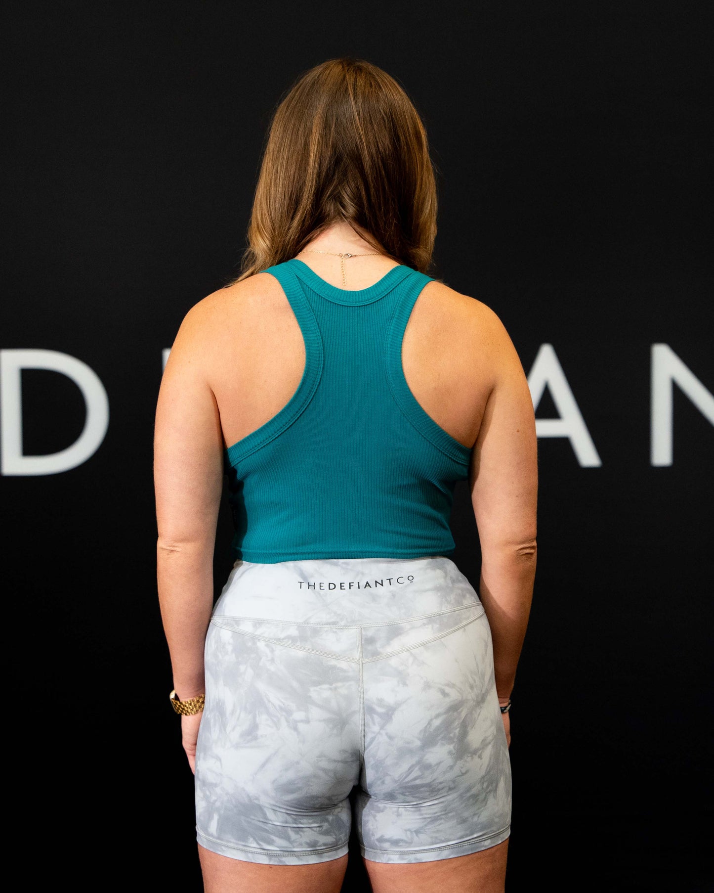 A girl against a black The Defiant Co backdrop wearing the DFNT. Ribbed Cropped Tank.  The tank is breathable with a stretchy fabric so is perfect for working out in.  It has a subtle DFNT. logo on a tag.  The colour is teal.