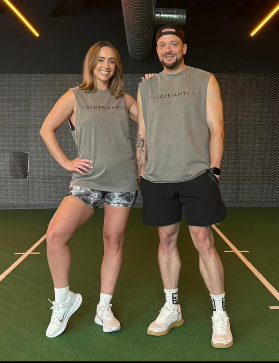 A photo showing a guy and girl wearing a unisex sleeveless t-shirt. The shirt has the famous ‘The Defiant Co’ logo across the front of the chest. The sleeveless has a round neck and is slightly oversized. The sleeveless is faded grey