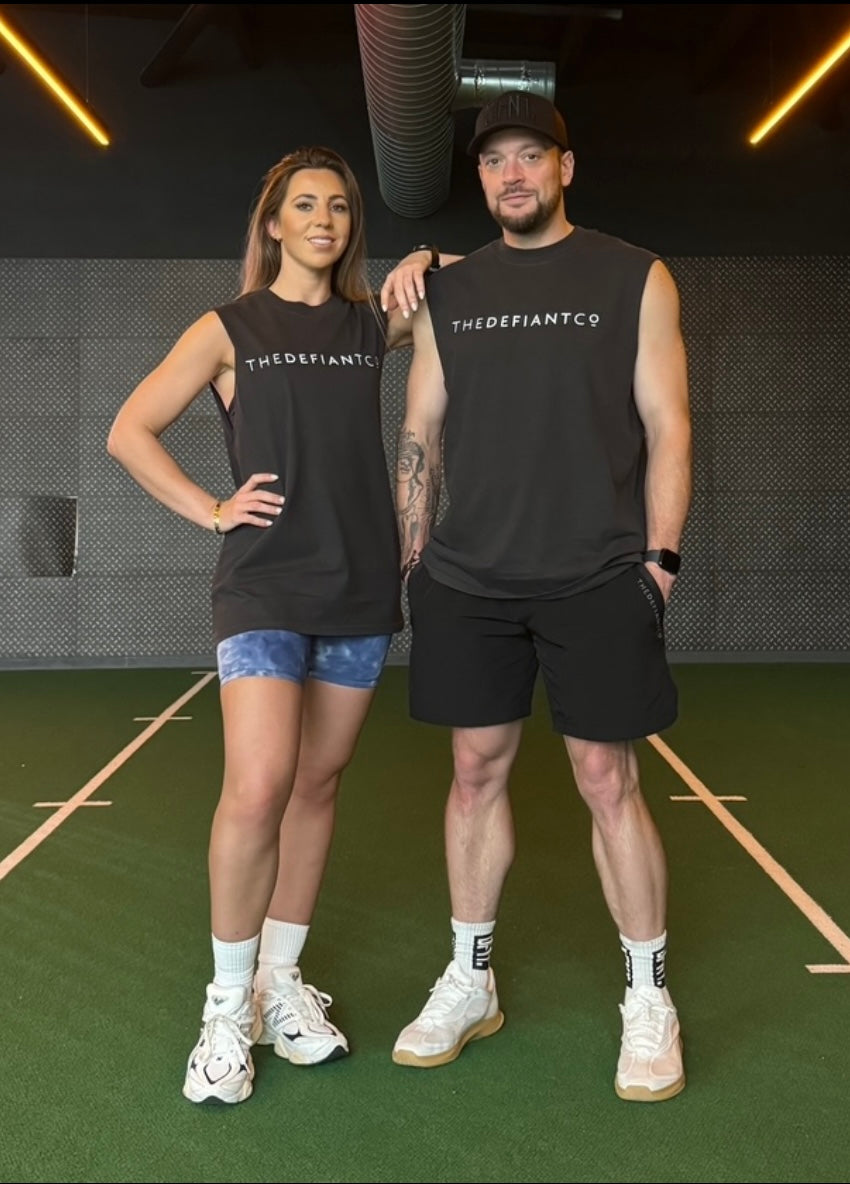 A photo showing a guy and girl wearing a unisex sleeveless t-shirt. The shirt has the famous ‘The Defiant Co’ logo across the front of the chest. The sleeveless has a round neck and is slightly oversized. The sleeveless is faded black..