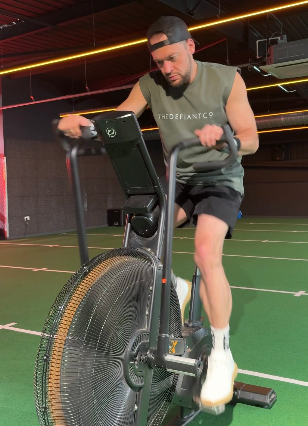 A photo showing a guy wearing a faded sleeveless t-shirt. The shirt has the famous ‘The Defiant Co’ logo across the front of the chest. The sleeveless has a round neck and is slightly oversized. The guy is performing an exercise on an air bike. The sleeveless is forest green.