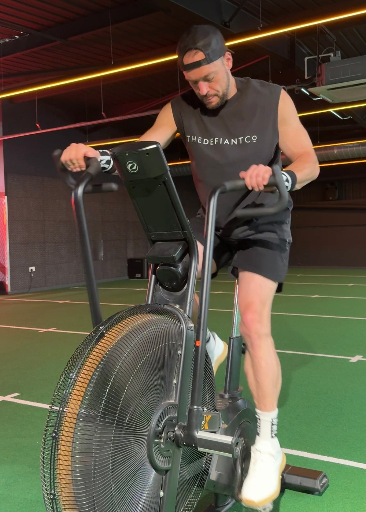 A photo showing a guy wearing a faded sleeveless t-shirt. The shirt has the famous ‘The Defiant Co’ logo across the front of the chest. The sleeveless has a round neck and is slightly oversized. The guy is performing an exercise on an air bike. The sleeveless is faded black..