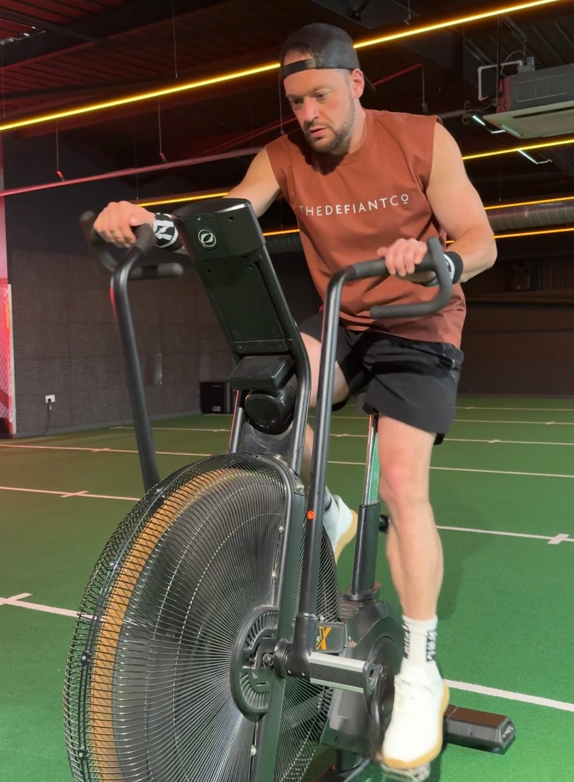 A photo showing a guy wearing a faded sleeveless t-shirt. The shirt has the famous ‘The Defiant Co’ logo across the front of the chest. The sleeveless has a round neck and is slightly oversized. The guy is performing an exercise on an air bike. The sleeveless is Burnt Orange.