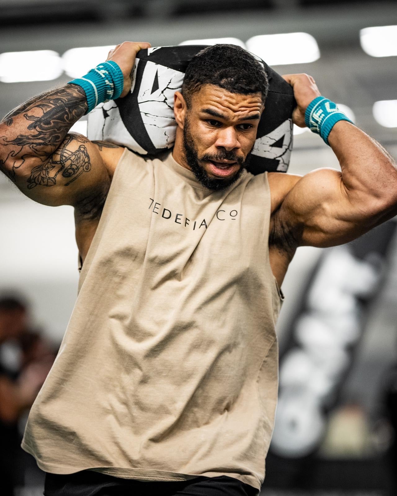 A photo showing a guy wearing a faded sleeveless t-shirt.  The shirt has the famous ‘The Defiant Co’ logo across the front of the chest.  The sleeveless has a round neck and is slightly oversized.  The sleeveless is faded khaki in colour.  The guys carrying a sandbag while running round a track in a crossfit workout.