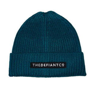 A photo showing a beanie hat. The hat has a ribbed finish with a roll cuff.  The cuff has The Defiant Co logo embroidered on the centre of the front in white on a black background. The beanie hat is teal.