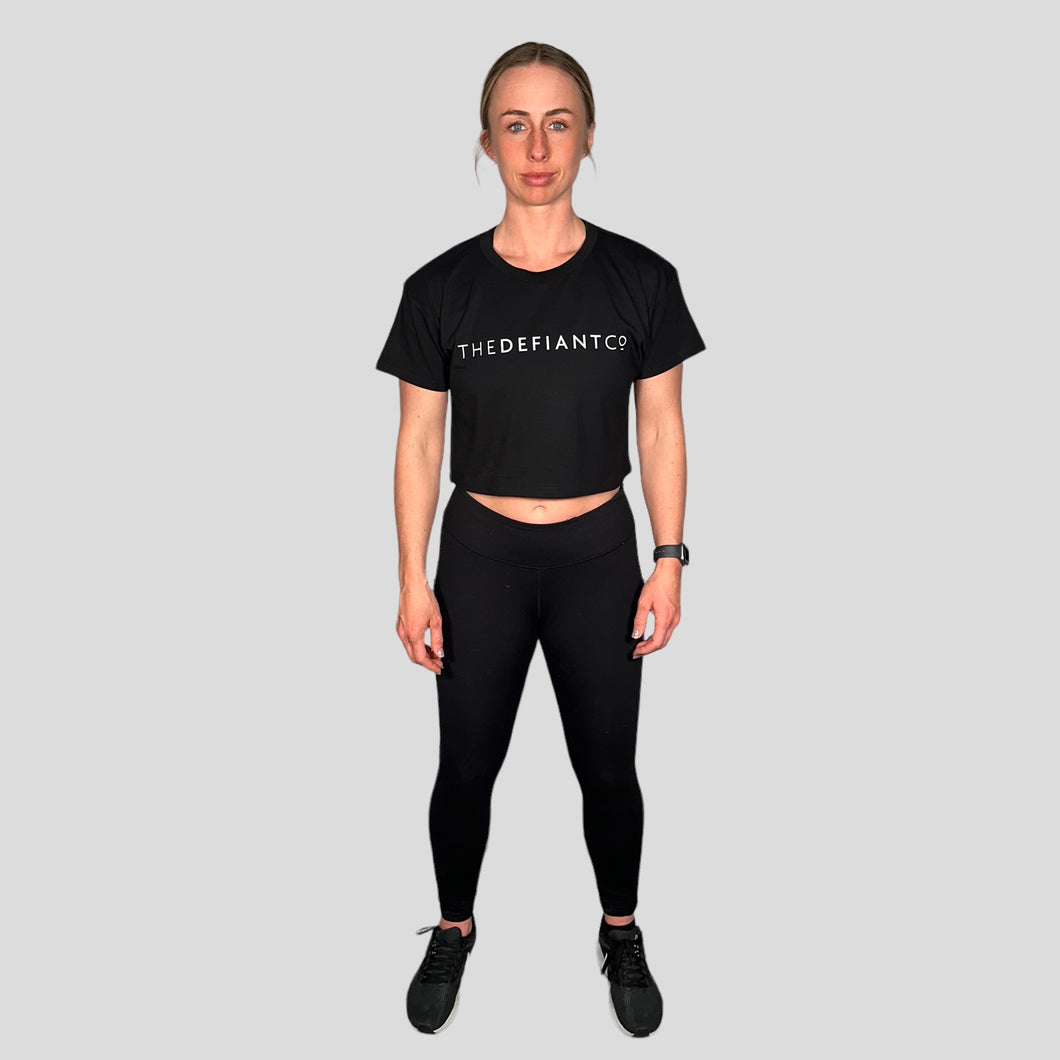 A girl wearing a cropped t-shirt .  The shirt is a female git and has a round neck and standard sleeve length.  The shirt has The Defiant Co logo across the centre of the chest.  The shirt is black in colour.