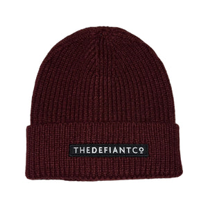 A photo showing a beanie hat. The hat has a ribbed finish with a roll cuff.  The cuff has The Defiant Co logo embroidered on the centre of the front in white on a black background. The beanie hat is burgundy.