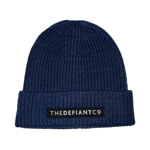 A photo showing a beanie hat. The hat has a ribbed finish with a roll cuff.  The cuff has The Defiant Co logo embroidered on the centre of the front in white on a black background. The beanie hat is steel blue.